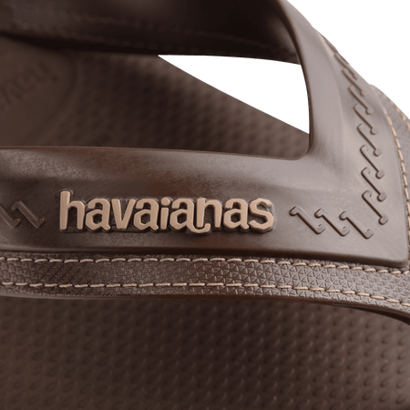 dark brown sandals with rice pattern and grooves, with brown straos with a lighter brown stitched-look detailing, zoomed in view of light brown havaianas logo on strap