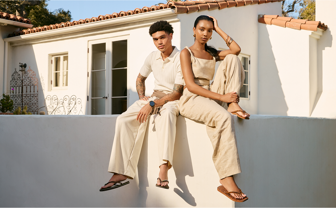 a man and a women sitting next to each other on a white wall. The men is wearing a brown top flip flop while the woman is wearing a slim square flip flop, the new velvet style