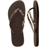 Pair of brown Havaianas flip flops with gold gems on the straps, on a white background with left shoe in focus