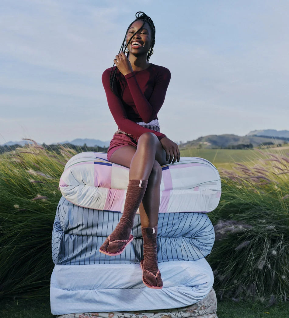 woman wearing socks with Havaianas flip flops sitting on a pile of mattresses in the middle of a grass field