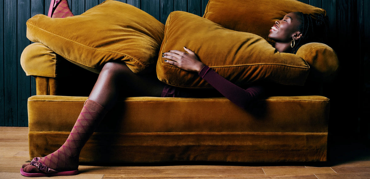 A woman reclines on a mustard-yellow velvet sofa wearing orange and mauve socks with magenta Havaianas flip flops, wrapped in matching cushions, against a dark paneled wall and wood floor.